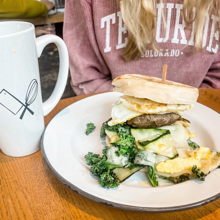 A large, stacked breakfast sandwich alongside a cup of hot coffee from The Butcher & The Baker