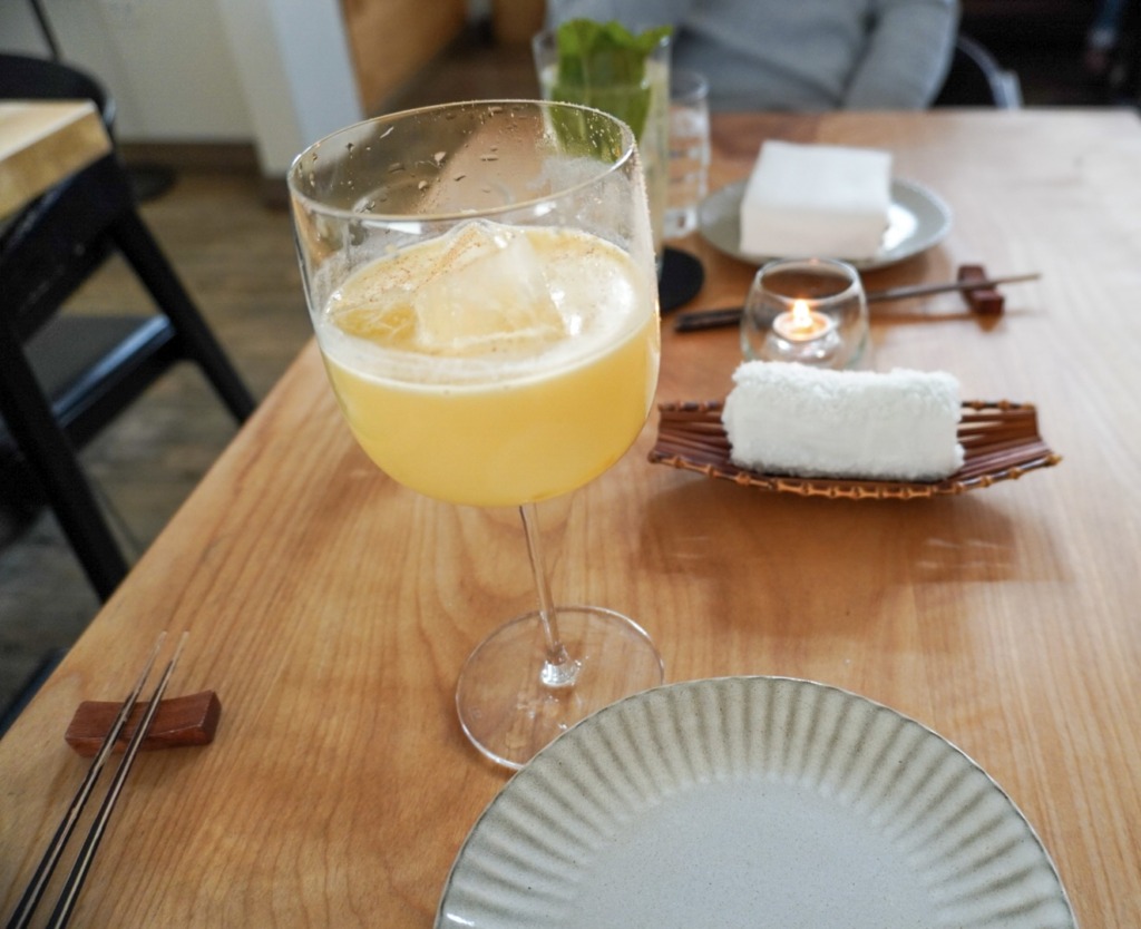 Golden cocktail with ice on Kumiko's minimalist wooden table set with chopsticks, plates, and a rolled towel.
