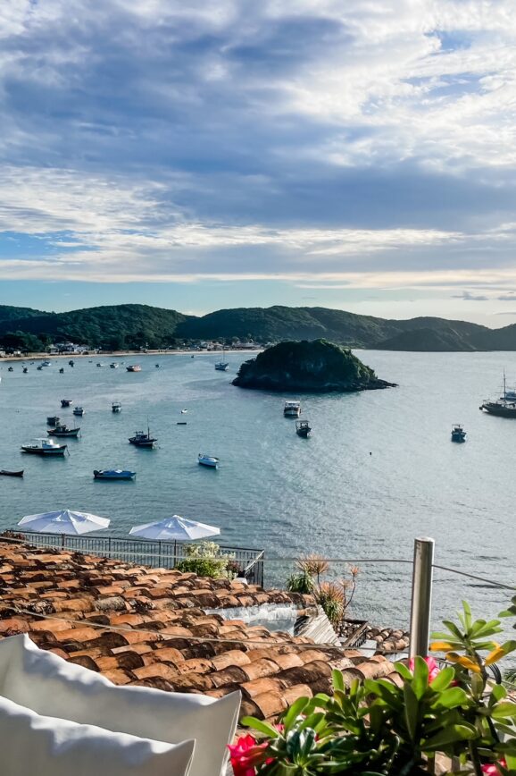 Scenic coastal view of Búzios, Brazil, with boats anchored in the bay and lush green hills under a partly cloudy sky.