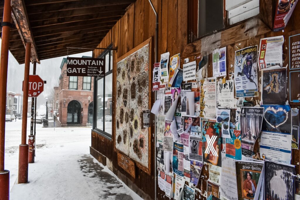 Snowy Telluride street corner with a wooden wall covered in colorful flyers and a sign pointing to Mountain Gate.