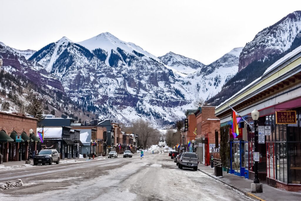 Snowy mountain backdrop over Telluride’s main street, lined with brick shops, colorful flags, and cozy charm.