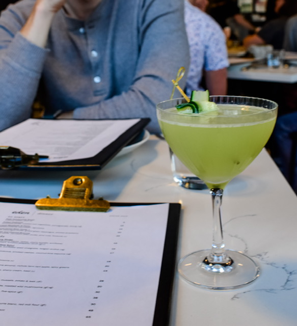 Bright green cucumber cocktail on a marble table beside menus at a modern restaurant.