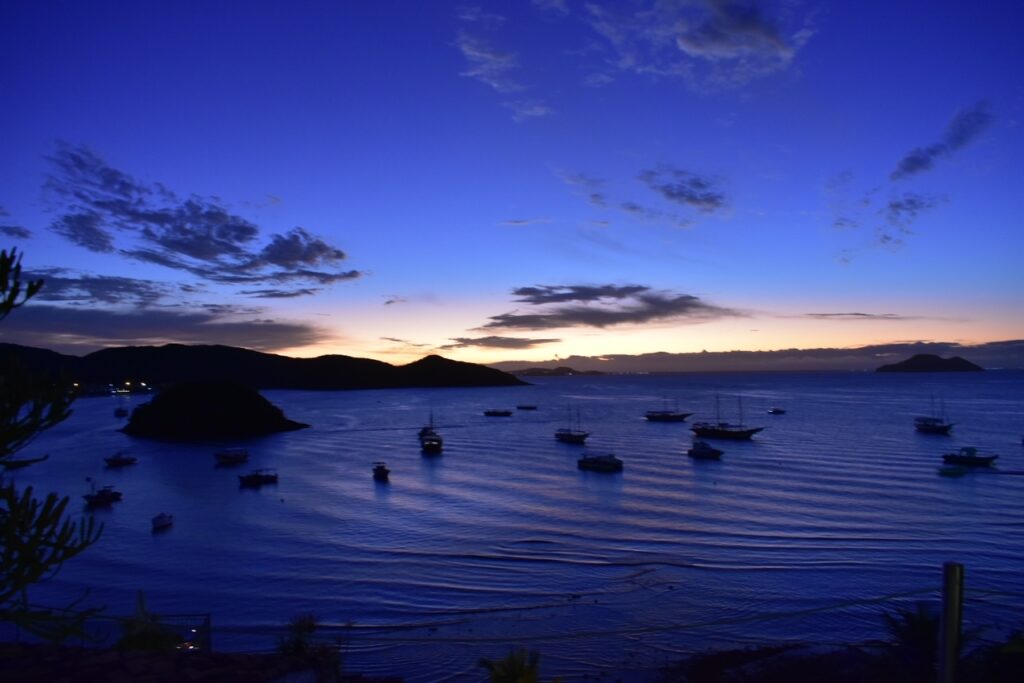 Indigo and sunset skies over Armação Bay in Búzios, Brazil, with boats floating on the calm water and lush green hills in the distance