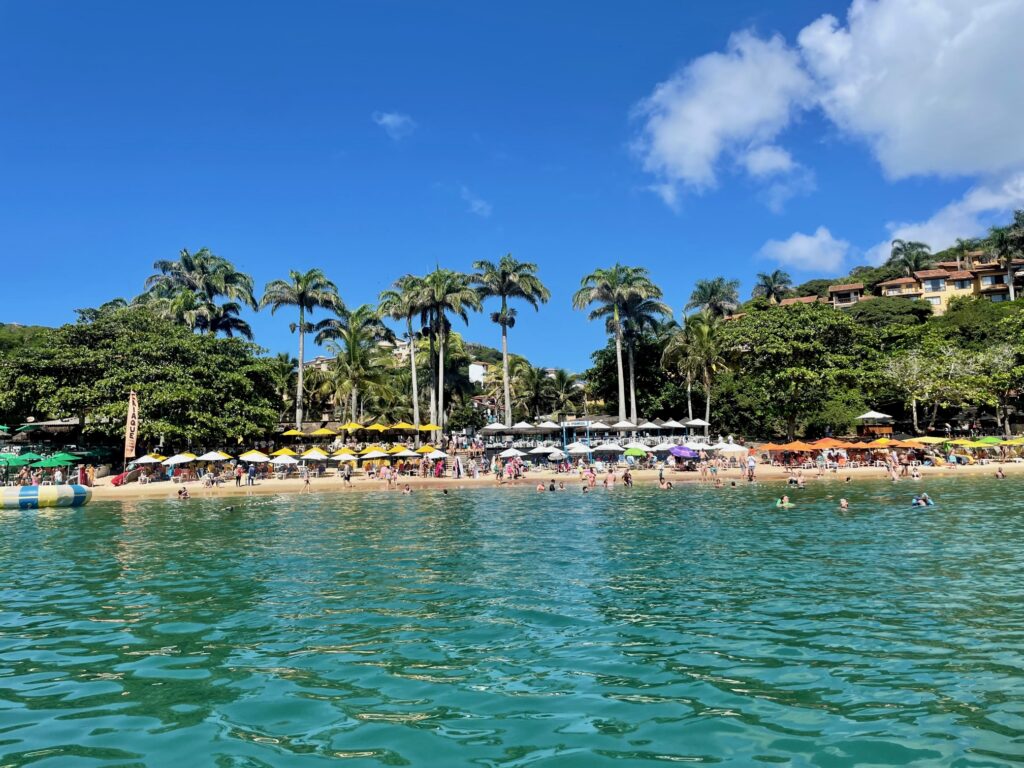 Palm-lined Joao Fernando Beach with calm waters and people relaxing under umbrellas.