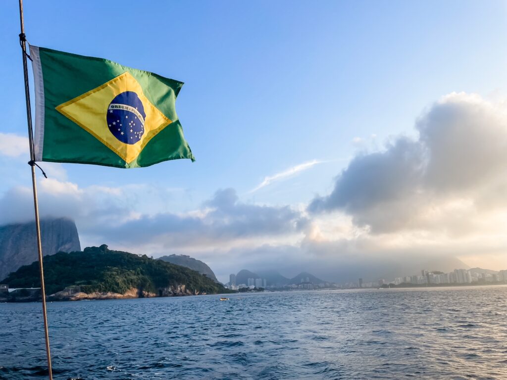 Brazilian flag waving above Guanabara Bay with Rio de Janeiro’s mountains and skyline in the distance.