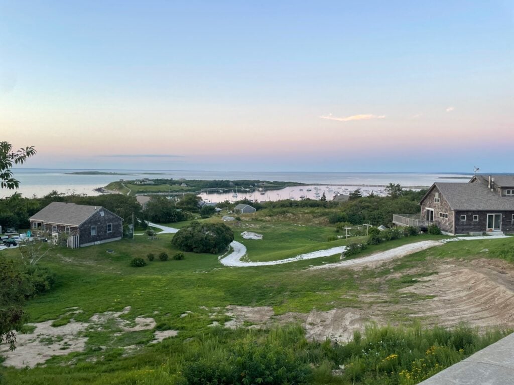 View of Cuttyhunk Island’s harbor at dusk with boats anchored offshore, grassy hills, and coastal cottages in the foreground.