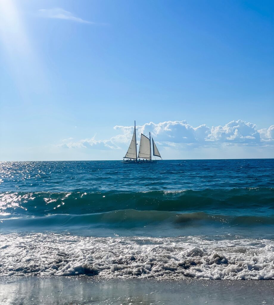 A sailboat glides across the ocean under a bright blue sky, viewed from the shore.