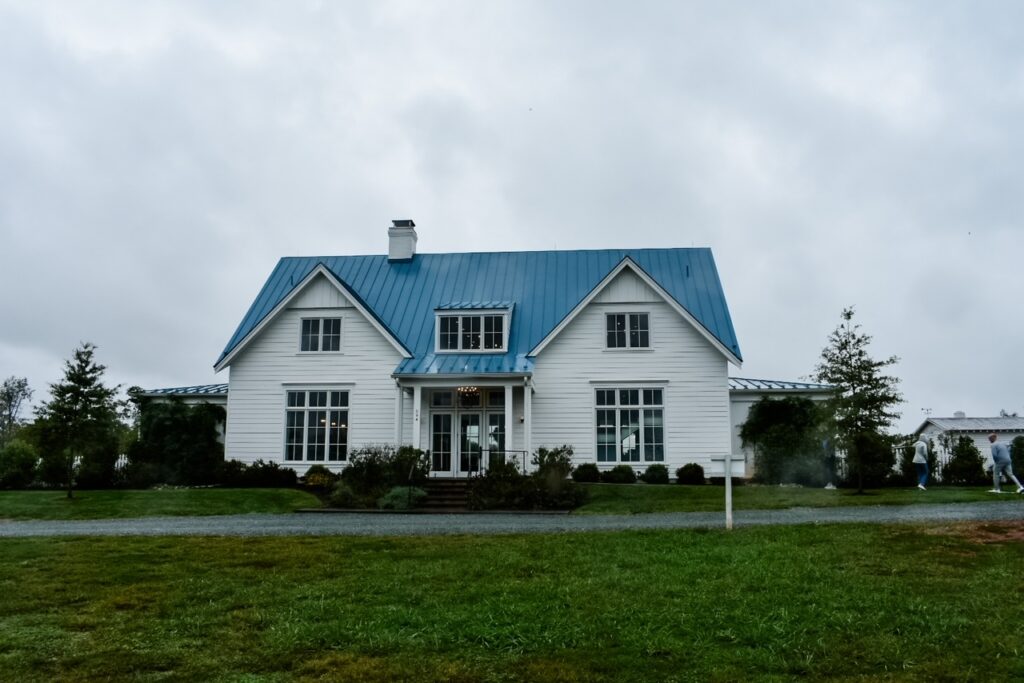 White farmhouse with a blue metal roof at Merrie Mill Farm & Vineyard on an overcast day.