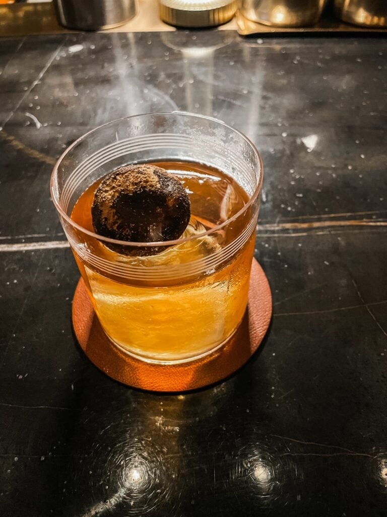 A smoky amber cocktail with a round garnish on an orange coaster.