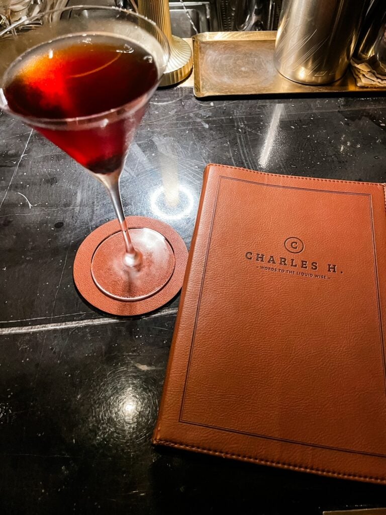 A red, cherry-infused Manhattan cocktail beside a leather-bound Charles H. menu on a dark bar counter.