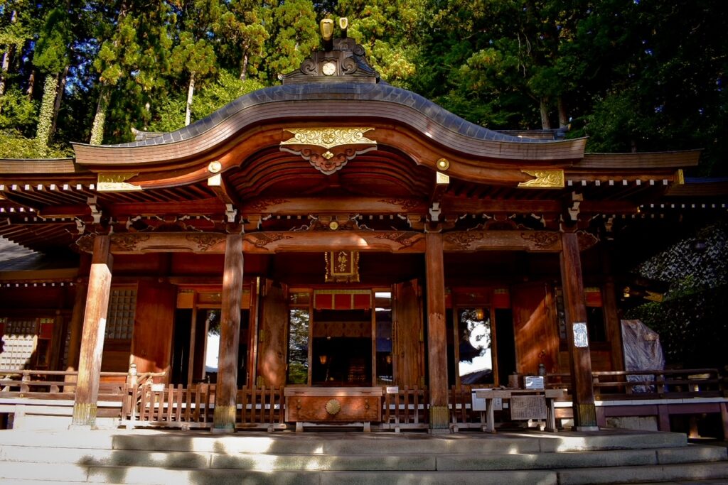 The ornate wooden main hall of Sakurayama Hachimangu Shrine, featuring intricate carvings, gold accents, and surrounded by tall trees.