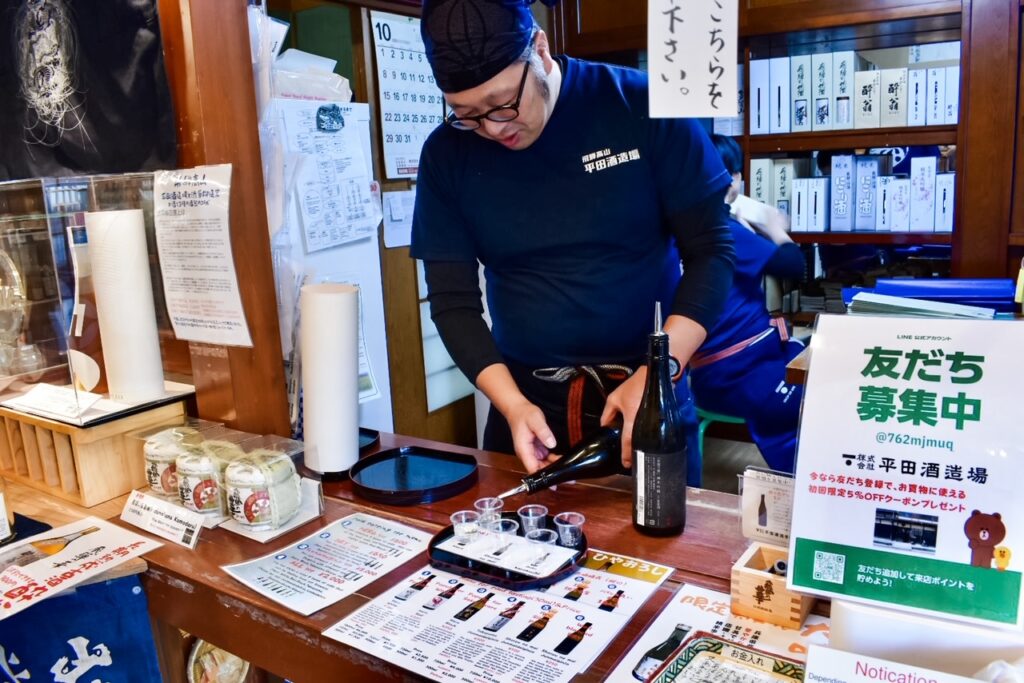 A staff member at a sake brewery pours samples at a wooden tasting counter surrounded by bottles, cups, and informational displays.