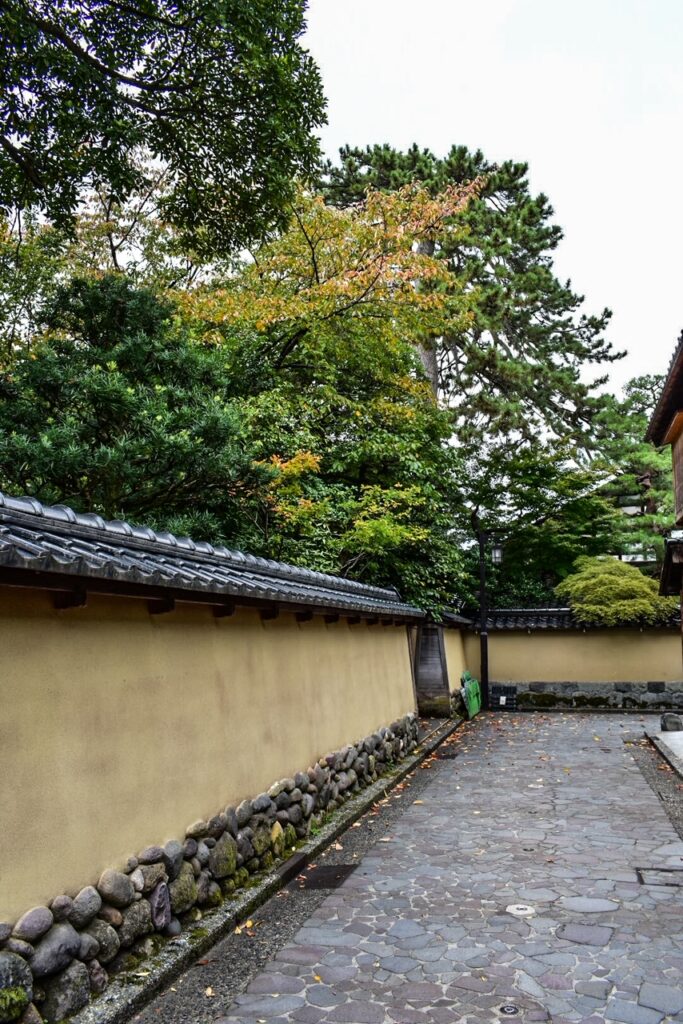 Narrow stone alley bordered by earthen walls and lush trees in Kanazawa’s historic Nagamachi samurai district.