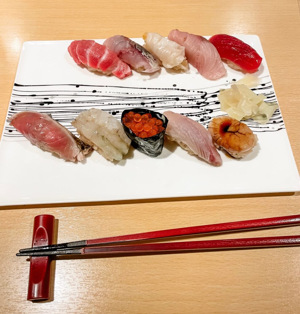 Assorted nigiri sushi, including tuna, shrimp, and salmon roe, elegantly arranged on a white plate with red chopsticks beside it.