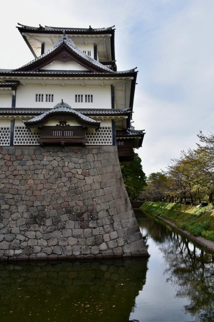 Traditional Japanese castle tower with white walls and tiled roofs rising above a stone base and moat in Kanazawa.