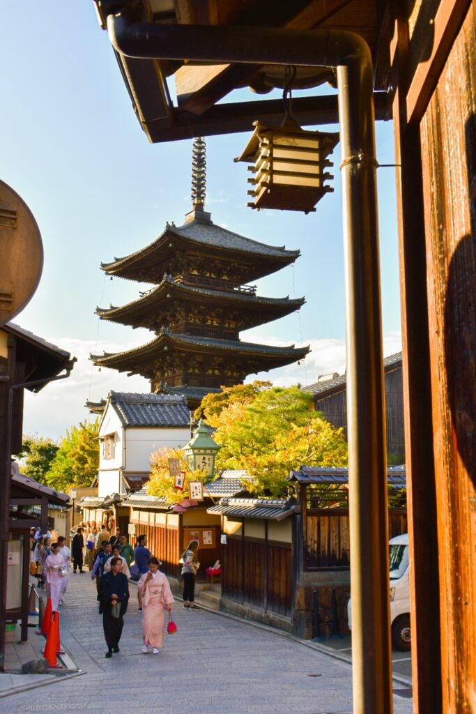 A scenic street view in Sannenzaka, Kyoto, with traditional wooden buildings leading toward the Yasaka Pagoda under a bright sky.
