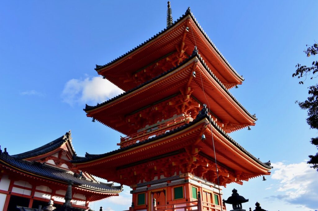 The vivid red and orange three-tiered pagoda of Kiyomizu-dera Temple in Kyoto, standing tall against a bright blue sky.