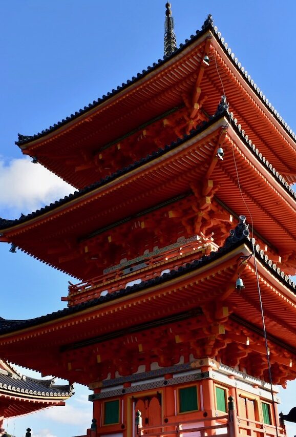 The vivid red and orange three-tiered pagoda of Kiyomizu-dera Temple in Kyoto, standing tall against a bright blue sky.
