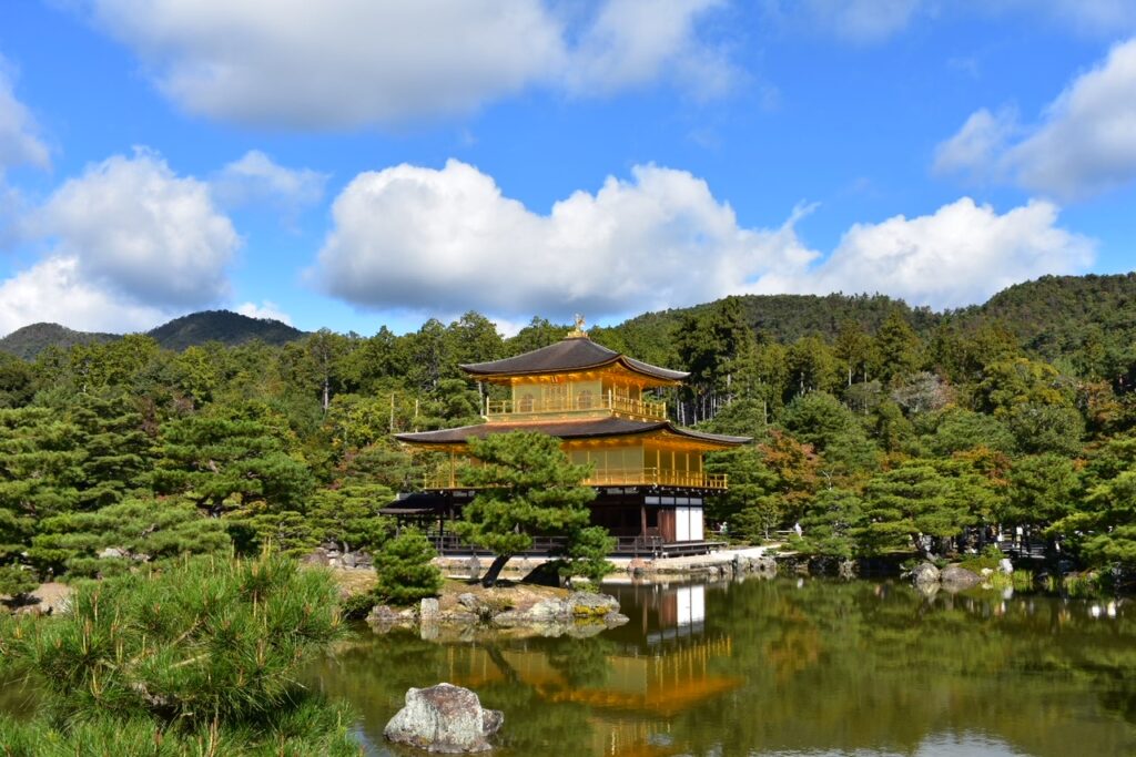 The iconic Kinkakuji, or Golden Pavilion, gleams in sunlight beside a serene reflective pond surrounded by lush green trees and distant mountains.