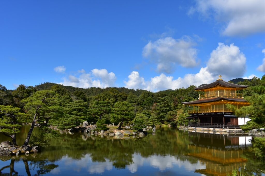 The Golden Pavilion (Kinkakuji) in Kyoto reflected on the still waters of its surrounding pond, framed by lush greenery and a bright blue sky with scattered clouds.