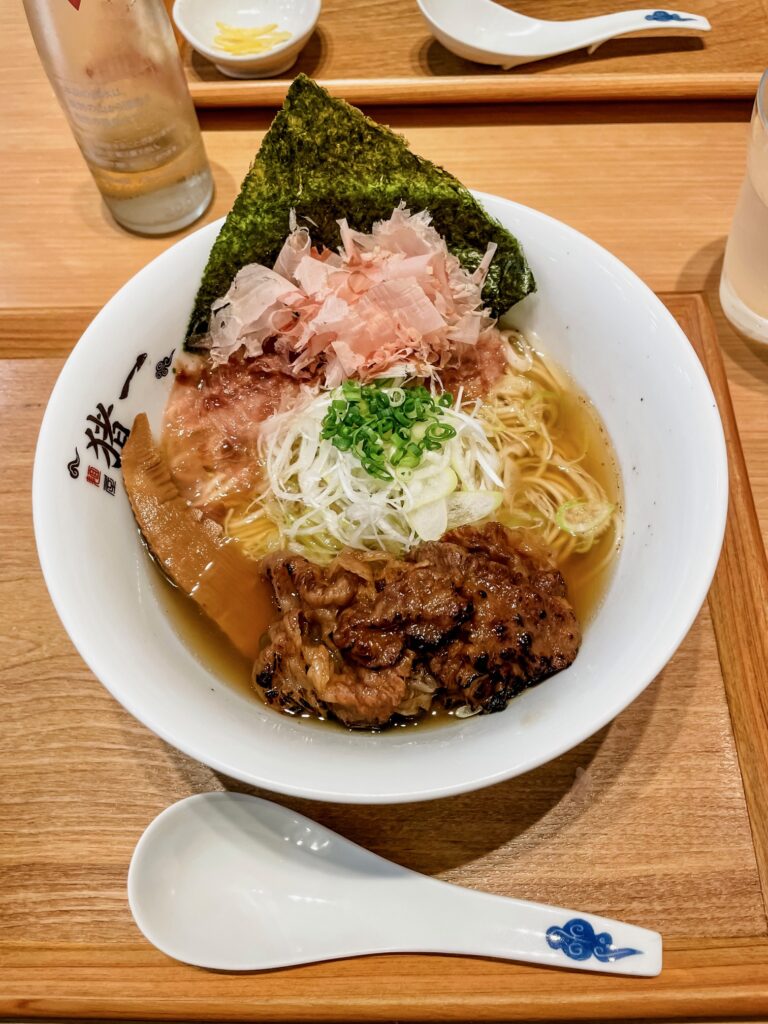 A beautifully presented bowl of ramen from Inoichi Ramen, featuring tender grilled meat, bamboo shoots, green onions, bonito flakes, and seaweed in a rich broth.