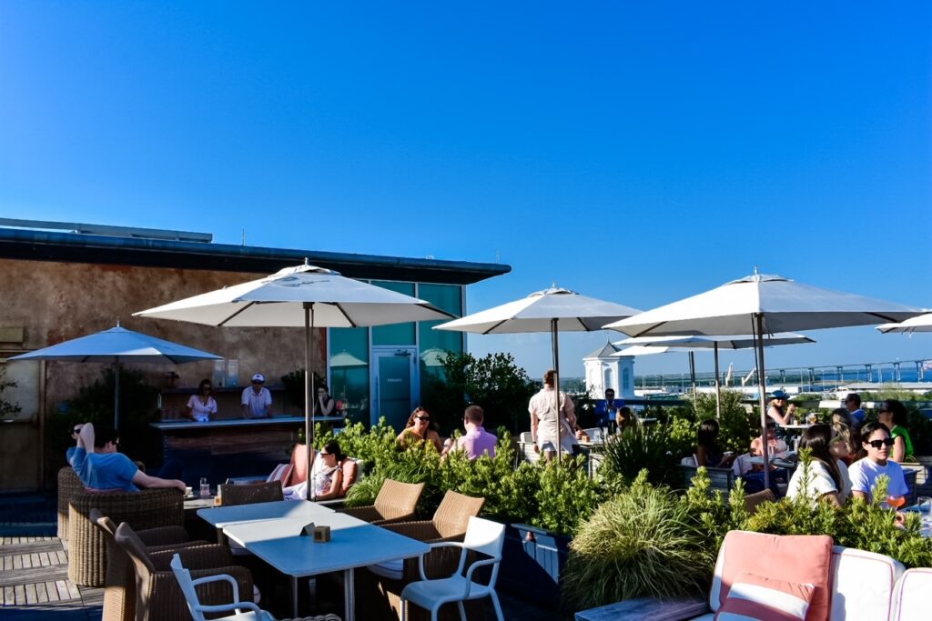 People relaxing under white umbrellas at Citrus Club's rooftop bar with ocean and bridge views in the background.