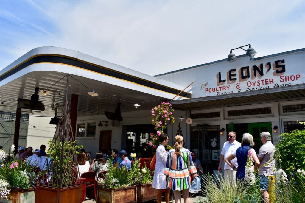 People gathered outside Leon’s Poultry & Oyster Shop in Charleston, enjoying a sunny day at the restaurant’s lively outdoor seating area surrounded by plants.