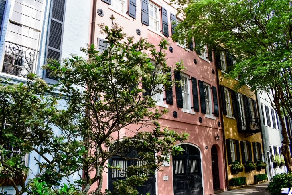 A row of pastel-colored historic houses on Rainbow Row in Charleston, with pink, yellow, and blue facades partially shaded by leafy trees.