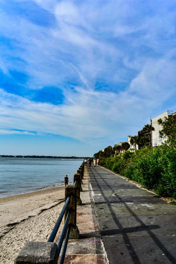 People walking along Charleston’s scenic waterfront promenade by the beach under a bright blue sky.