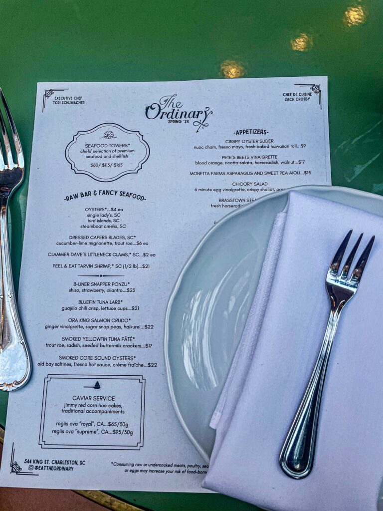 A close-up of a dining setup at The Ordinary restaurant in Charleston, featuring a neatly folded napkin with silverware on a plate beside the seafood-focused spring menu