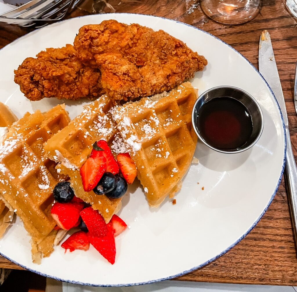 A plate of crispy fried chicken served with golden waffles topped with powdered sugar, fresh strawberries, and blueberries, accompanied by a small cup of maple syrup.