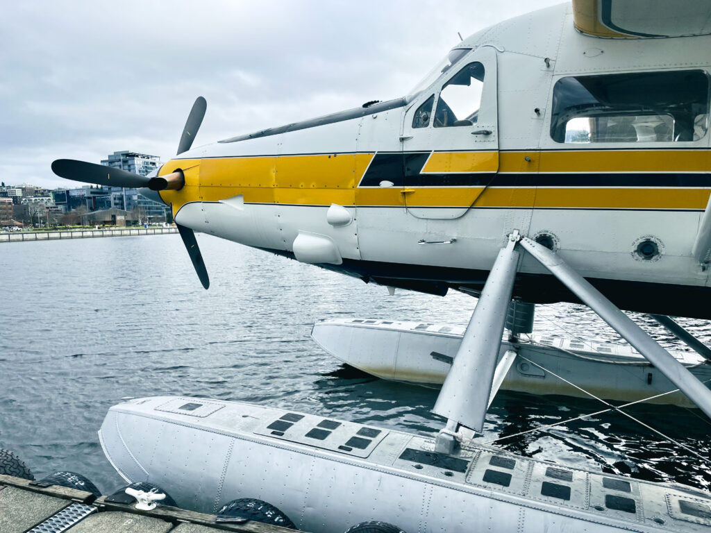 Yellow and white seaplane docked on calm water with city buildings in the background.