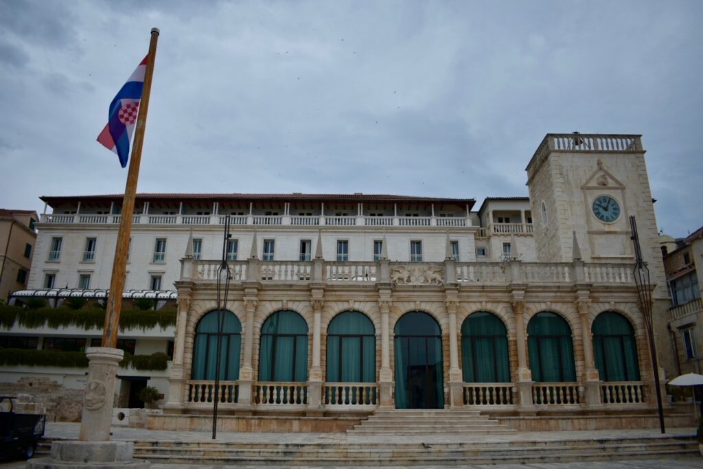 Palace Elisabeth in Hvar, Croatia, featuring grand arches, a clock tower, and the Croatian flag on a cloudy day.