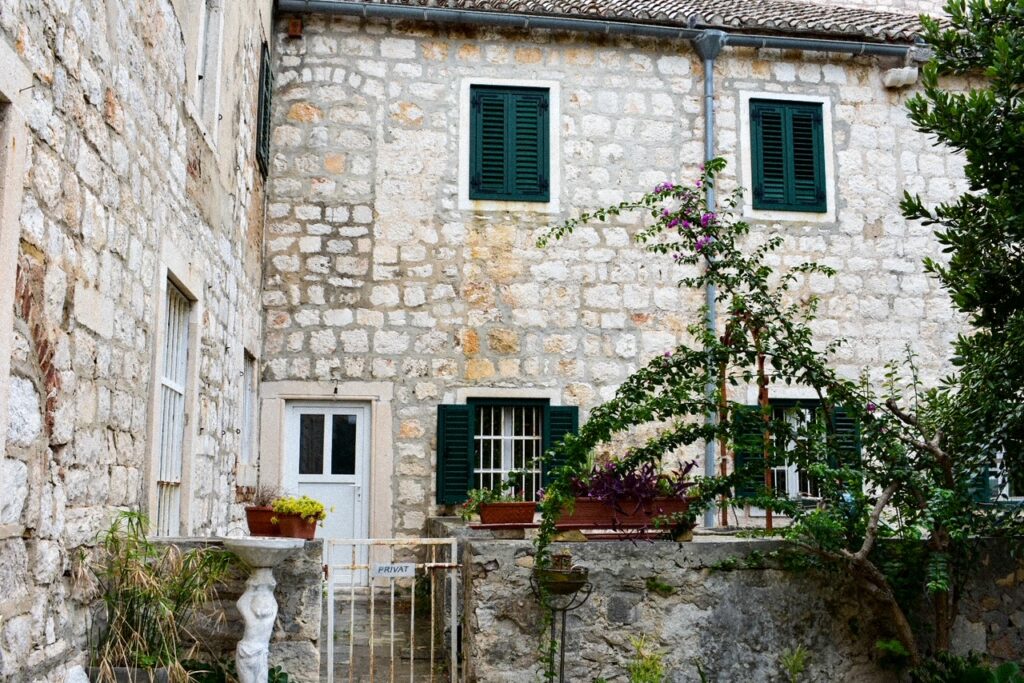 Charming stone courtyard with green shutters, potted plants, and flowering vines climbing the wall in Croatia.