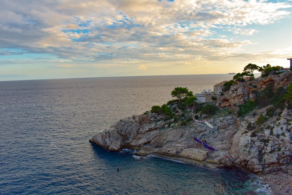 Rocky coastline with trees overlooking the calm sea at sunset under a cloudy sky.
