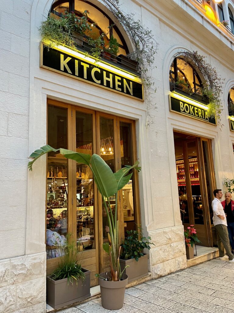Elegant exterior of Bokeria Kitchen in Split, Croatia, with tall plants and glowing yellow signage.