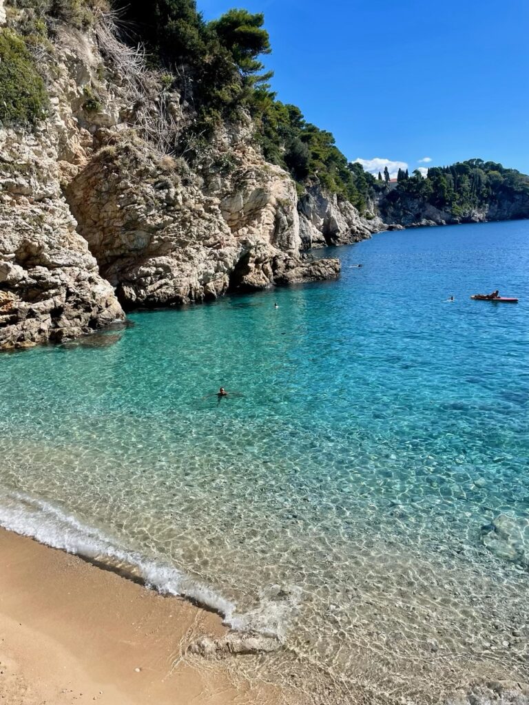 Crystal-clear turquoise waters lap against the sandy shore of Bellevue Beach, framed by rocky cliffs and greenery.
