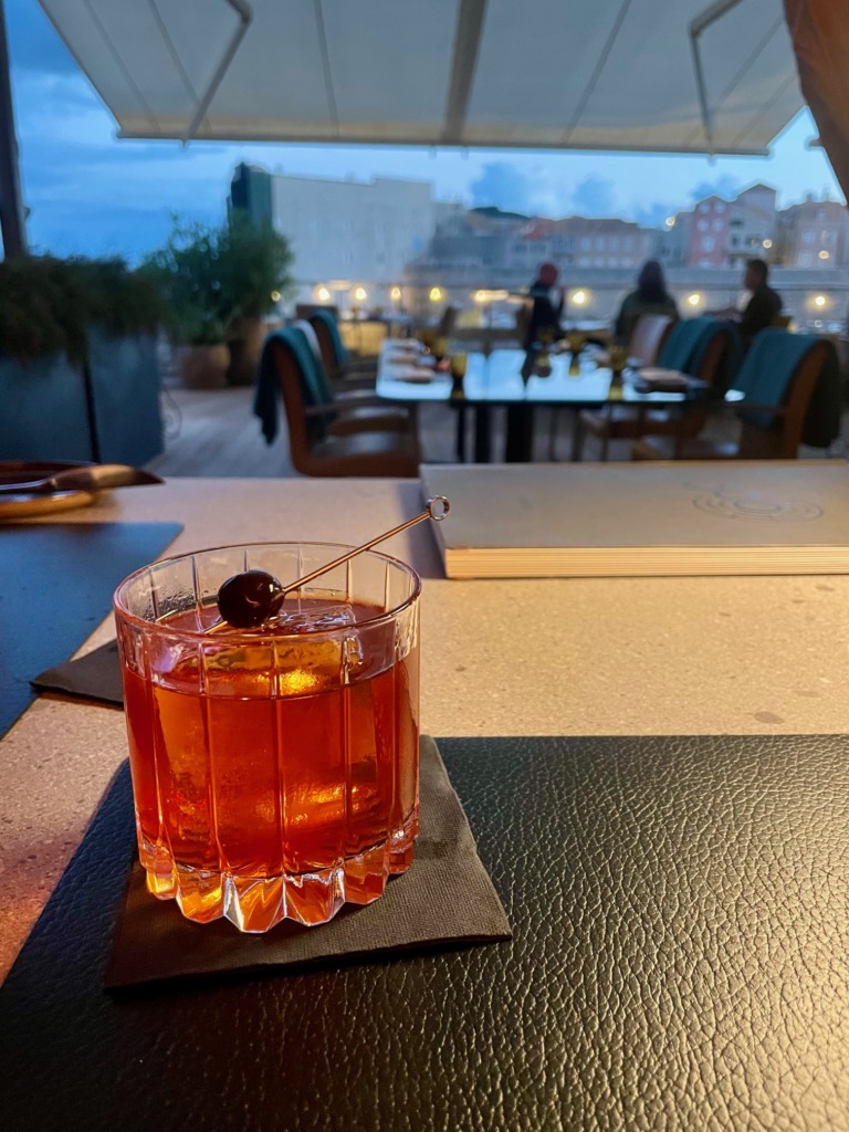 Amber cocktail garnished with cherries at a rooftop bar overlooking the city at dusk.