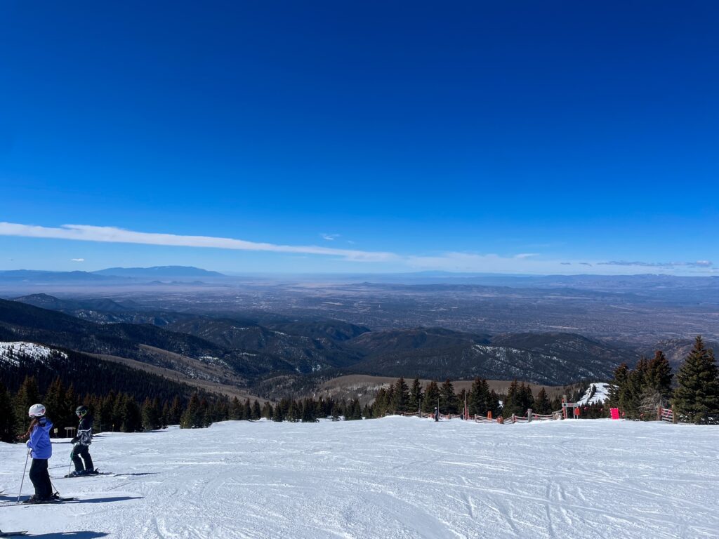 Skiers pause atop a snowy mountain slope, overlooking vast valleys and distant peaks under a vivid blue sky.