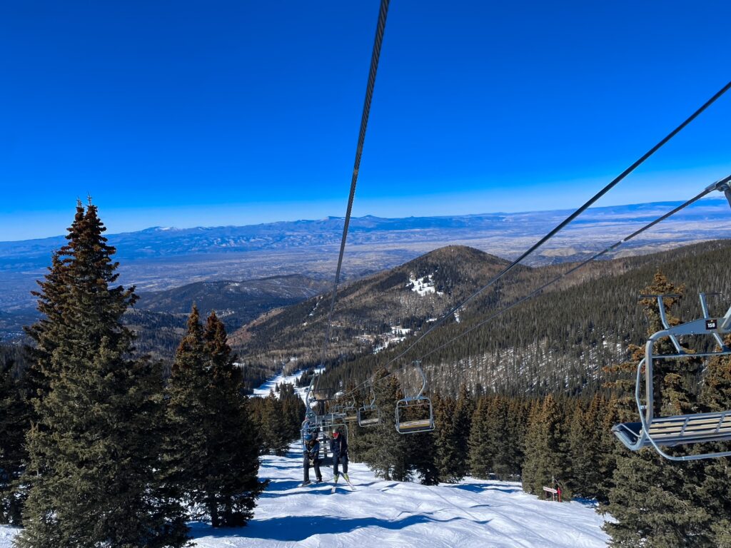 Skiers ride a lift above snow-covered slopes surrounded by pine trees under a brilliant blue mountain sky.