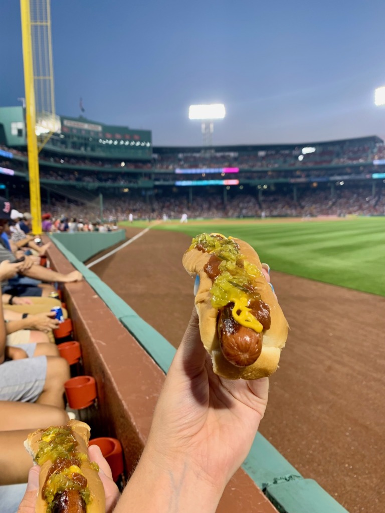 Hot dog with relish and mustard held up at a summer night Red Sox game at Fenway Park in Boston, Massachusetts, with the green field and packed stadium in the background