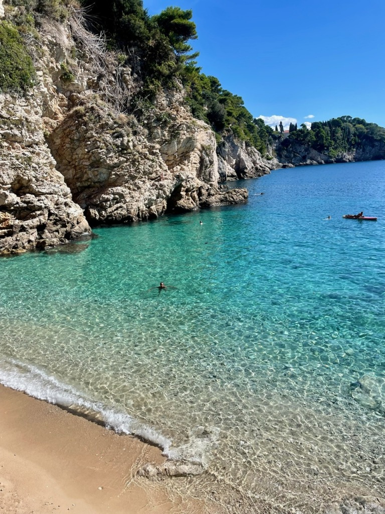 Crystal-clear turquoise water and a sandy cove along the Dalmatian Coast in Croatia, with limestone cliffs, pine trees, and swimmers enjoying the Adriatic Sea on a summer day