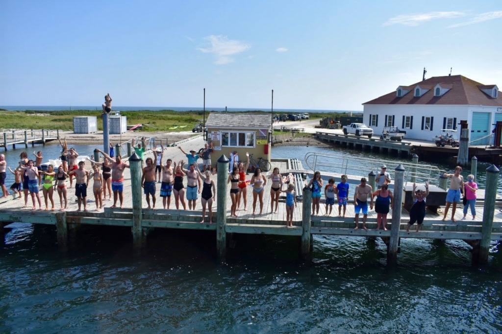 Group of summer vacationers in swimsuits cheering on a wooden dock at Cuttyhunk Island, Massachusetts, with the open ocean and a white harbor building behind them