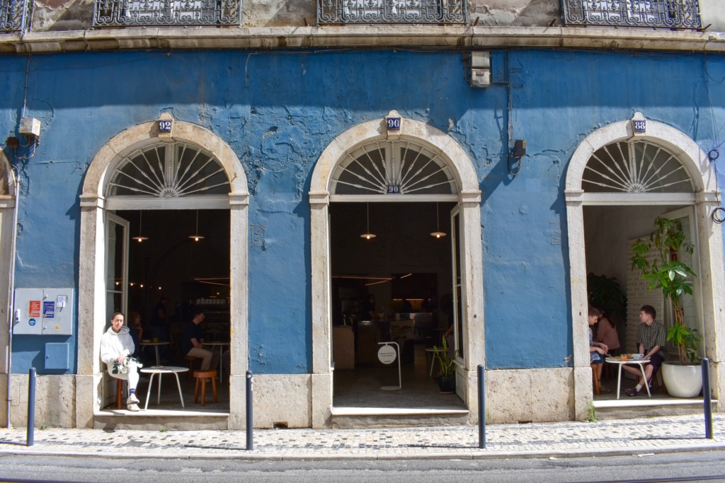 A blue building facade in Lisbon with three arched doorways opening into a cafe, people seated at small tables in the entryways