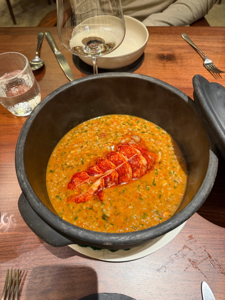 Lobster rice served in a black clay pot at Cozinha das Flores in Porto, Portugal — rich coral-orange broth with a whole lobster tail, with a wine glass alongside
