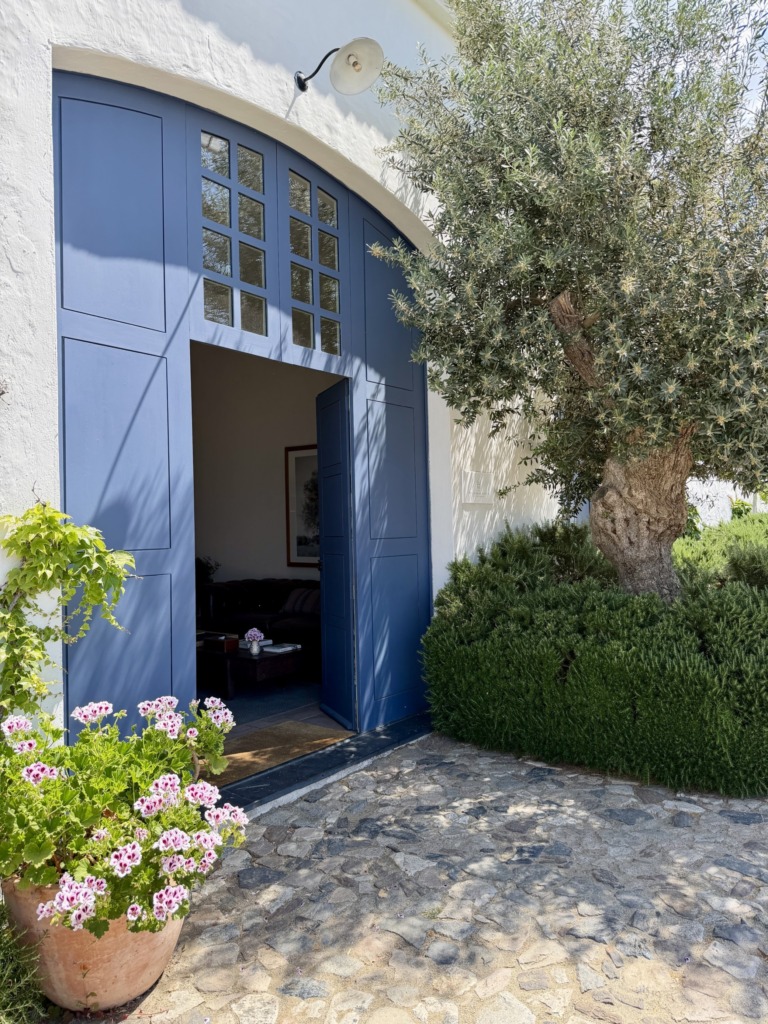 A large blue arched doorway opening into a guest room, flanked by a mature olive tree, pink geraniums in a terracotta pot, and a cobblestone path