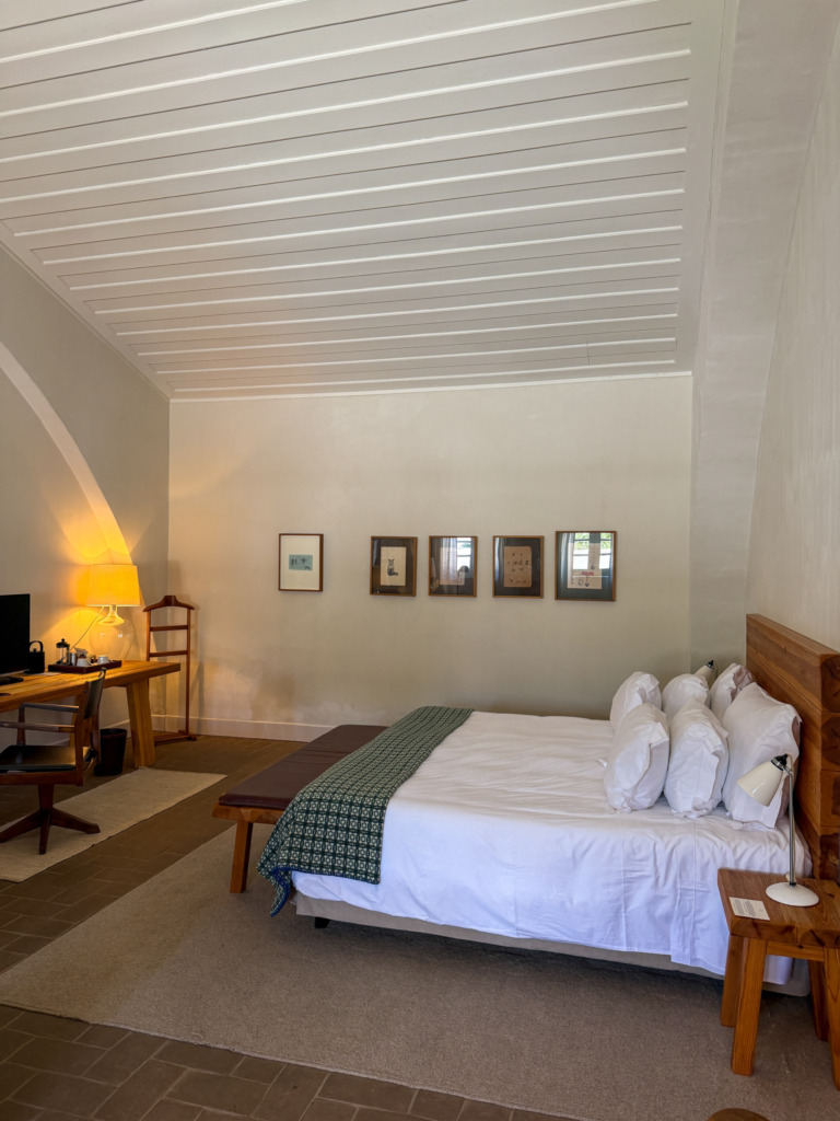 A king bed with a wooden headboard and green woven throw beneath a high vaulted ceiling, with rust-colored floor tiles and framed artwork lining the wall