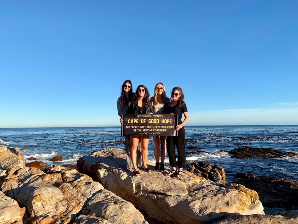 Four friends posing together at the Cape of Good Hope in Cape Town, holding the wooden sign marking the southernmost point, ocean and rocky cliffs behind them.