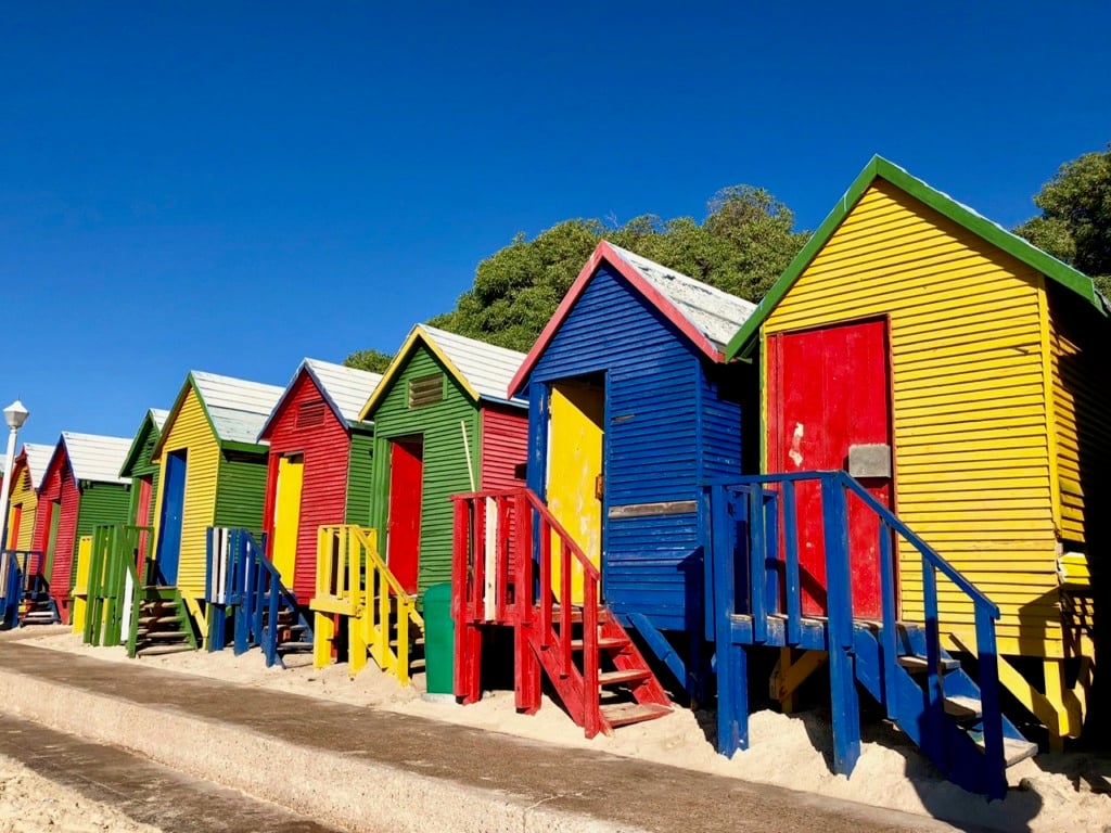 A row of brightly painted wooden beach huts in primary colors—red, yellow, blue, and green—with wooden stairs, sandy beach and palm trees behind them.