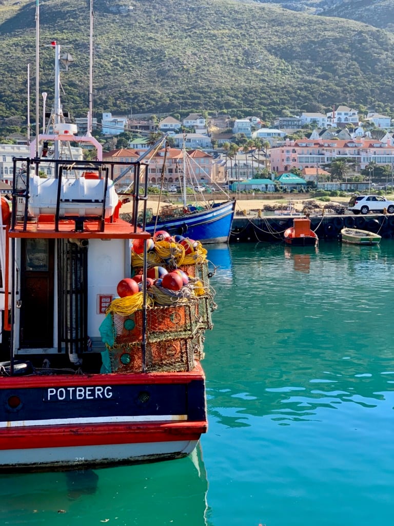 A red and white fishing boat moored in turquoise harbor water in Kalk Bay, with yellow and red buoys stacked on deck and colorful buildings climbing the hillside behind.